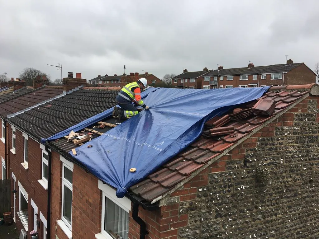 A roofer securing a heavy blue tarpaulin over storm-damaged roof tiles on a terraced house, grey sky