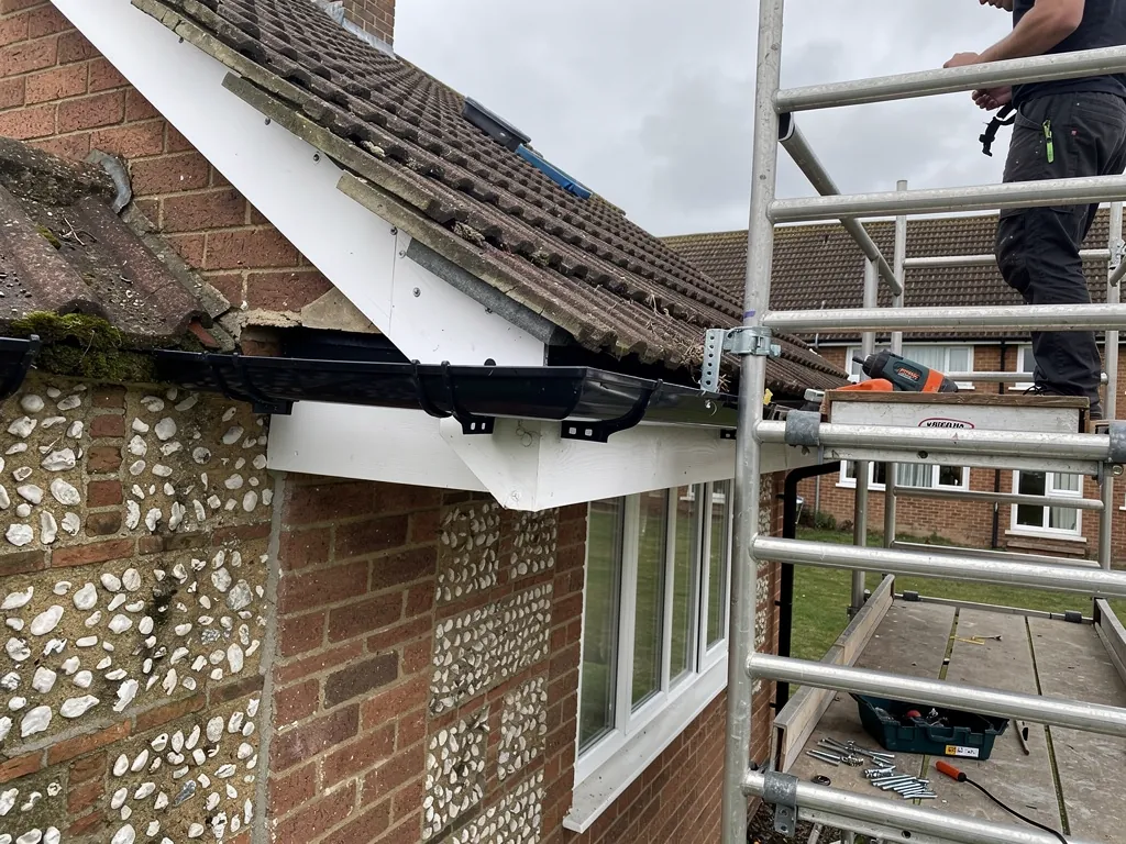 A roofer fitting a new gutter bracket and gutter run along a fascia board from a tower