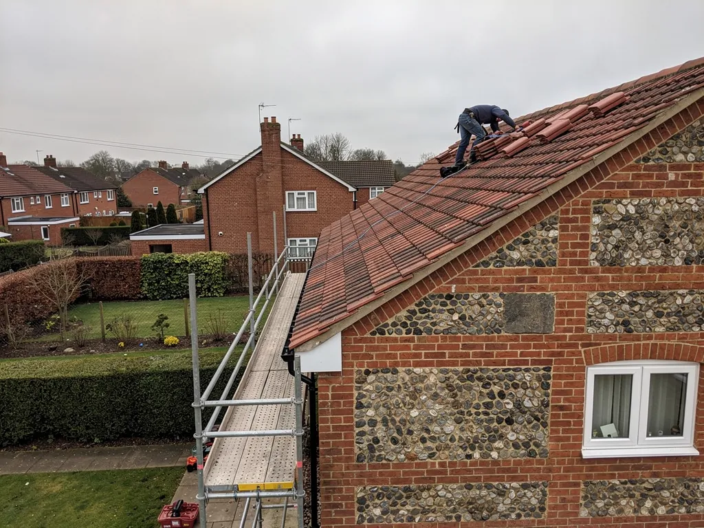 A roofer fixing new clay plain tiles on a pitched roof of a red-brick Hampshire house, scaffold at the eaves